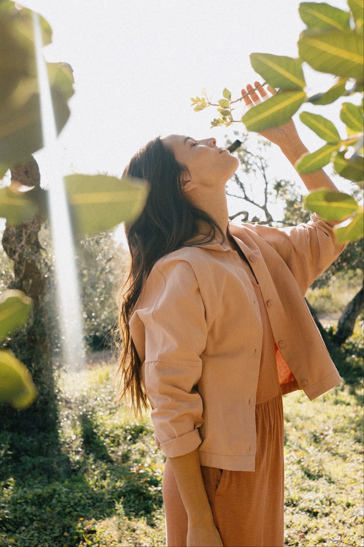 A woman in a light peach button-up shirt and matching peach skirt stands outdoors in a sunny garden, looking upward toward leaves while wearing a black crossbody bag.