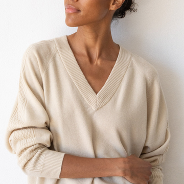A woman wearing a cream-colored V-neck knit sweater with textured cable detailing on the sleeves, standing against a plain white wall.