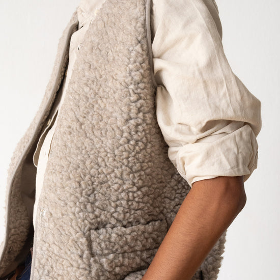 A close-up of a person wearing a light taupe sherpa fleece vest over a cream linen shirt, photographed against a clean white studio background.