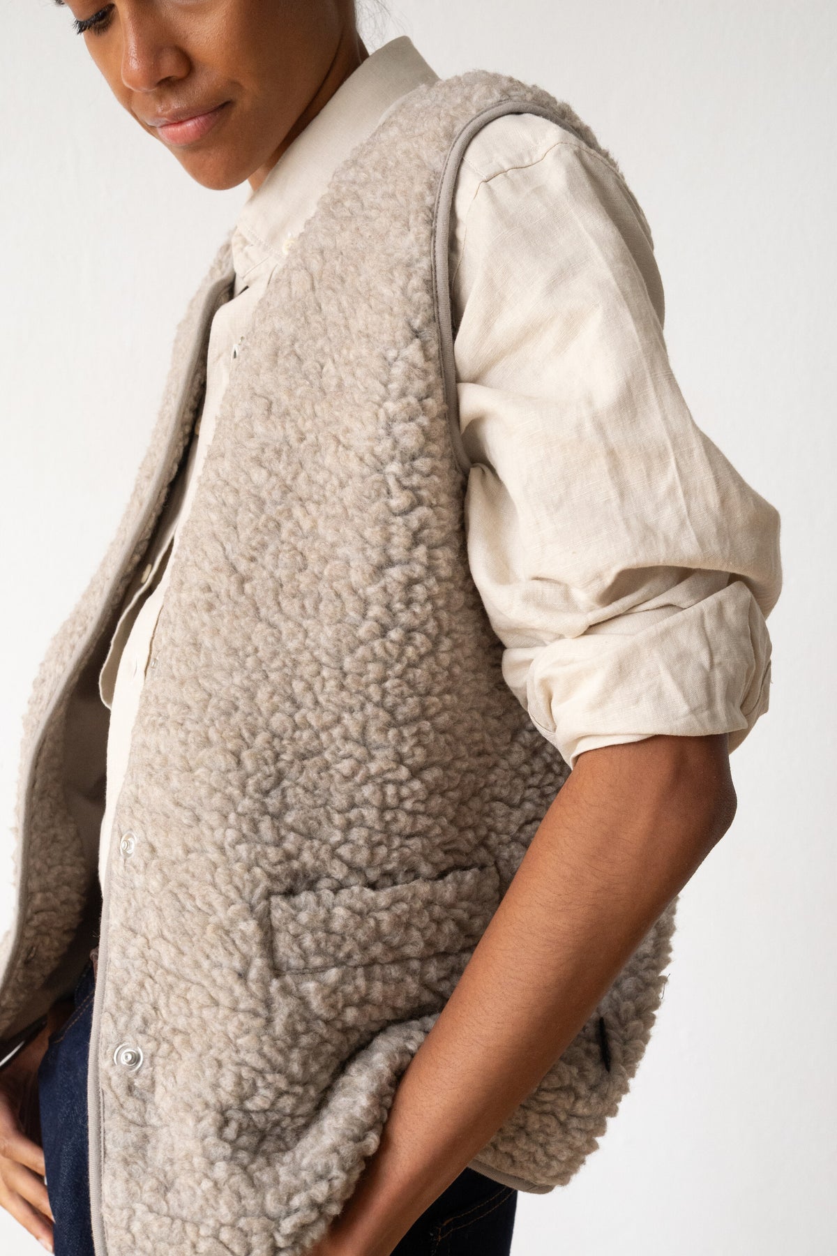 A close-up of a person wearing a light taupe sherpa fleece vest over a cream linen shirt, photographed against a clean white studio background.