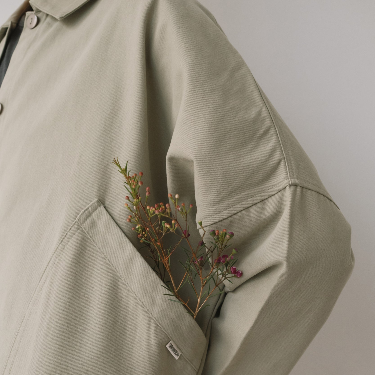Person wearing a green jacket with a pocket holding dried flowers against a plain background