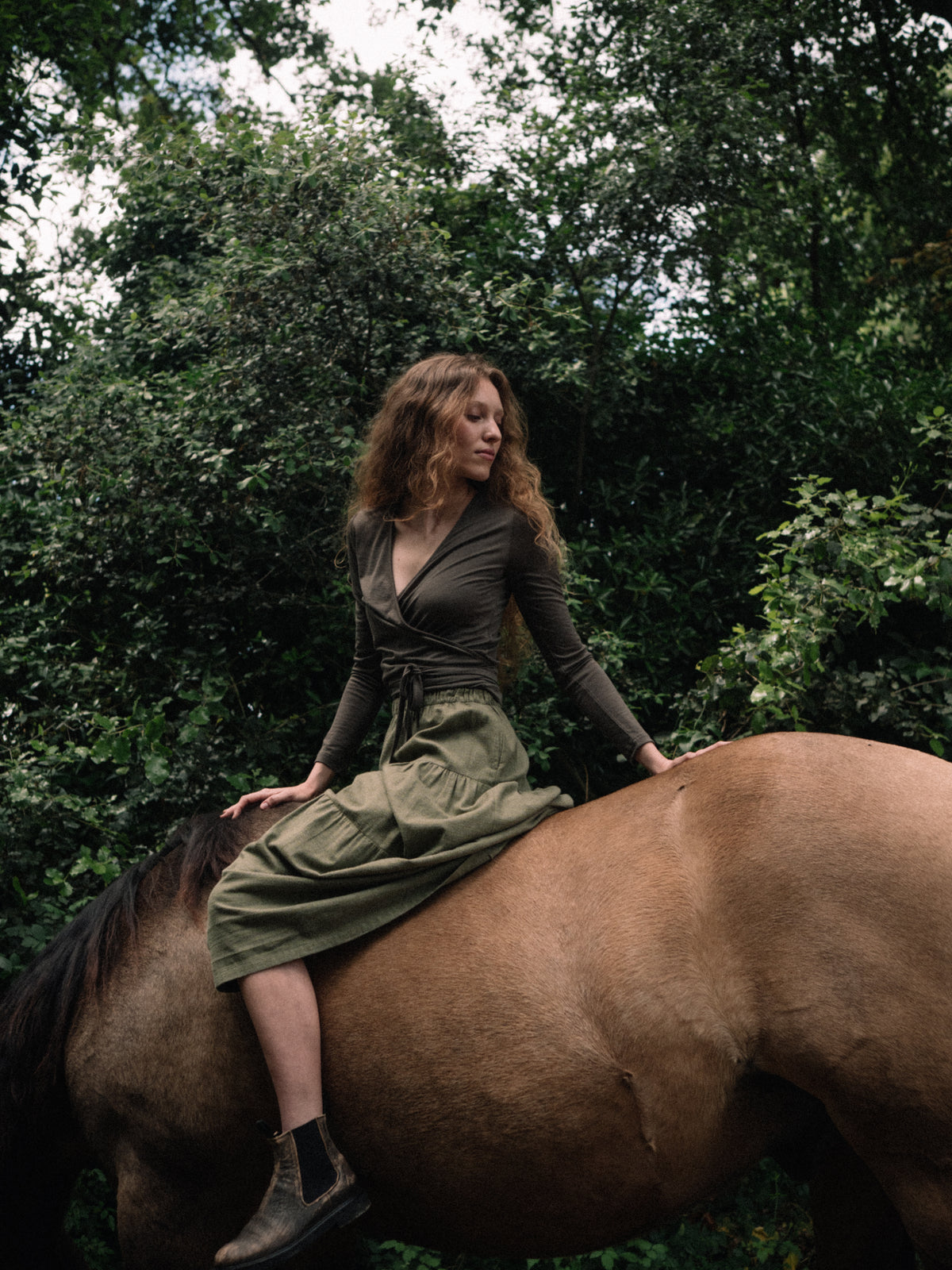 A woman wearing an olive green wrap dress and boots sits on a brown horse in a dense green forest setting.