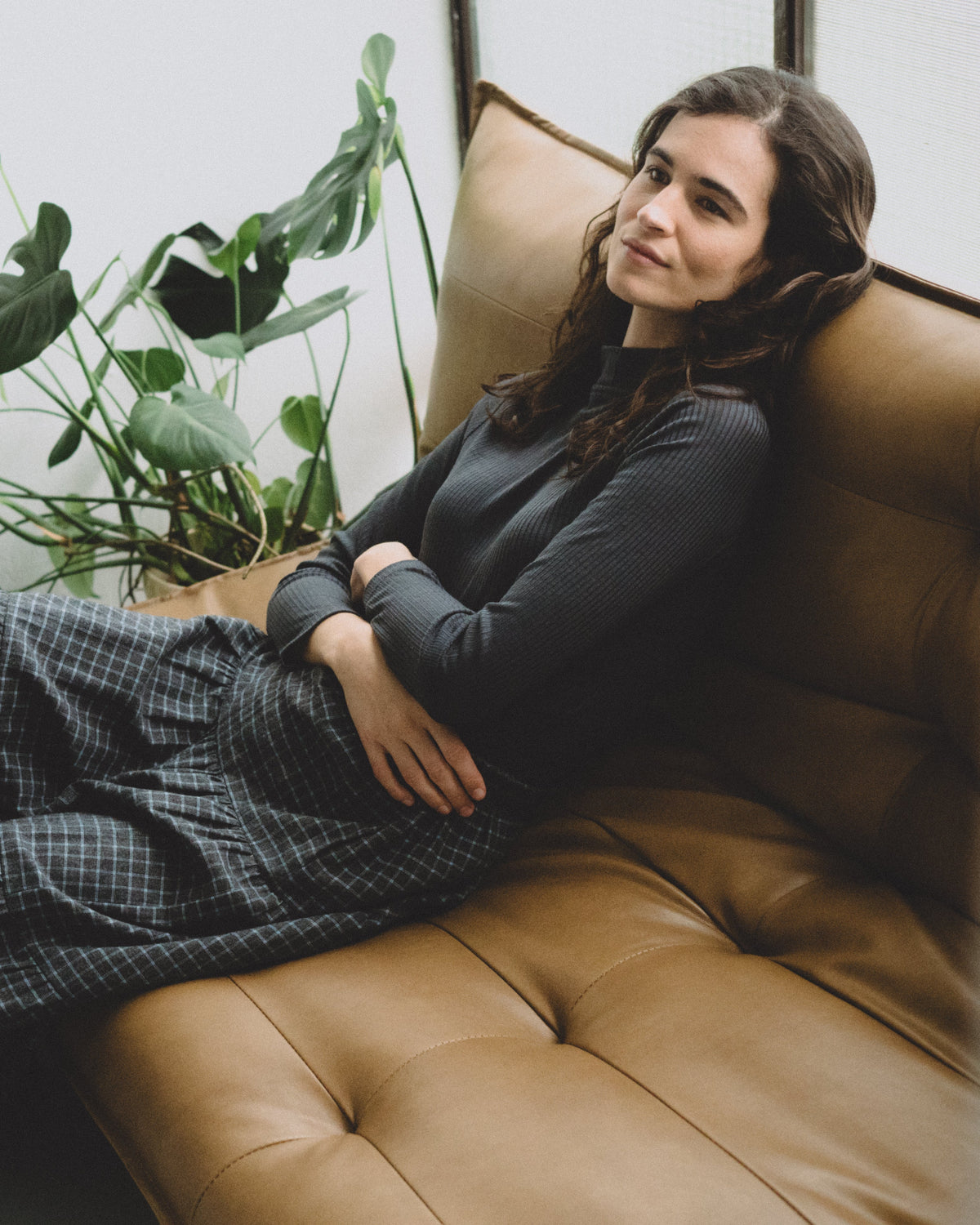 A woman reclining on a tufted brown leather sofa next to a green potted plant, wearing a dark charcoal long-sleeve top and a dark blue and white checkered skirt.