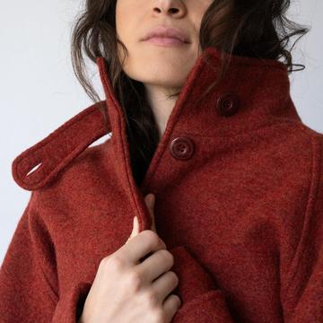 Close-up of a person wearing a textured rust-red wool coat with a high buttoned collar and dark buttons, holding the collar open with one hand against a plain light background.