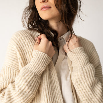 A close-up of a woman wearing a cream-colored ribbed knit cardigan over a light beige button-up shirt, set against a plain light background.